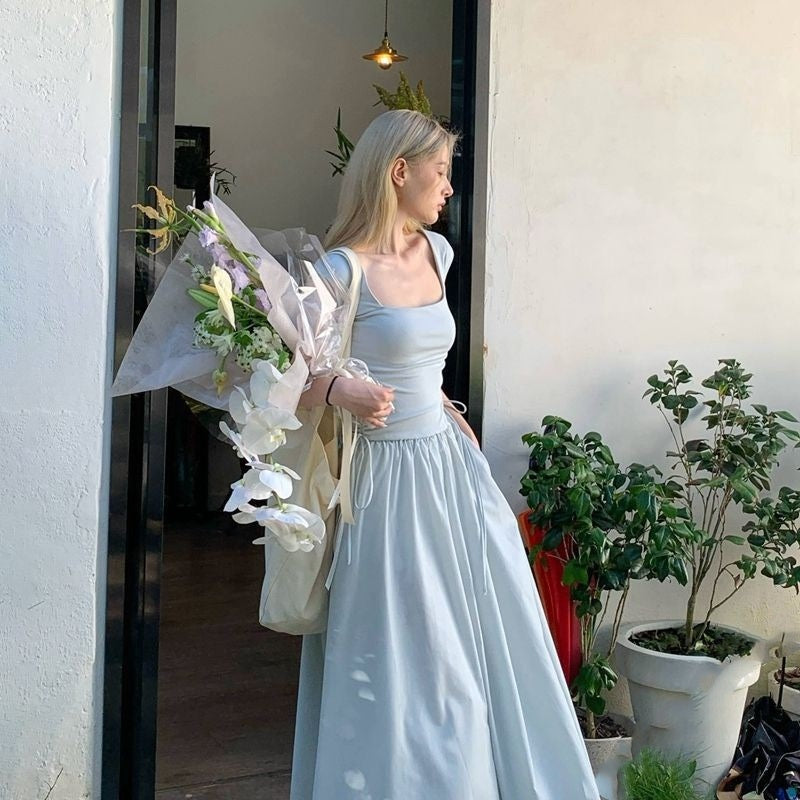 Woman in a light blue dress holding flowers, standing in front of a white wall with plants.
